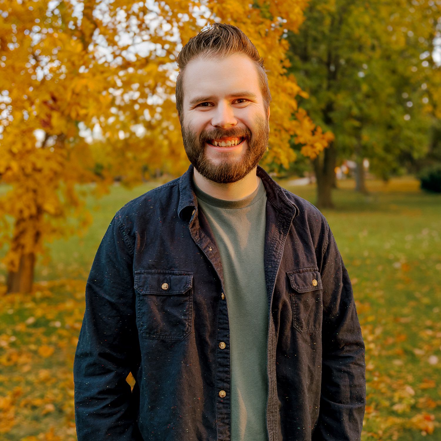 A smiling man with a beard in a speckled shirt stands among yellow autumn leaves.
